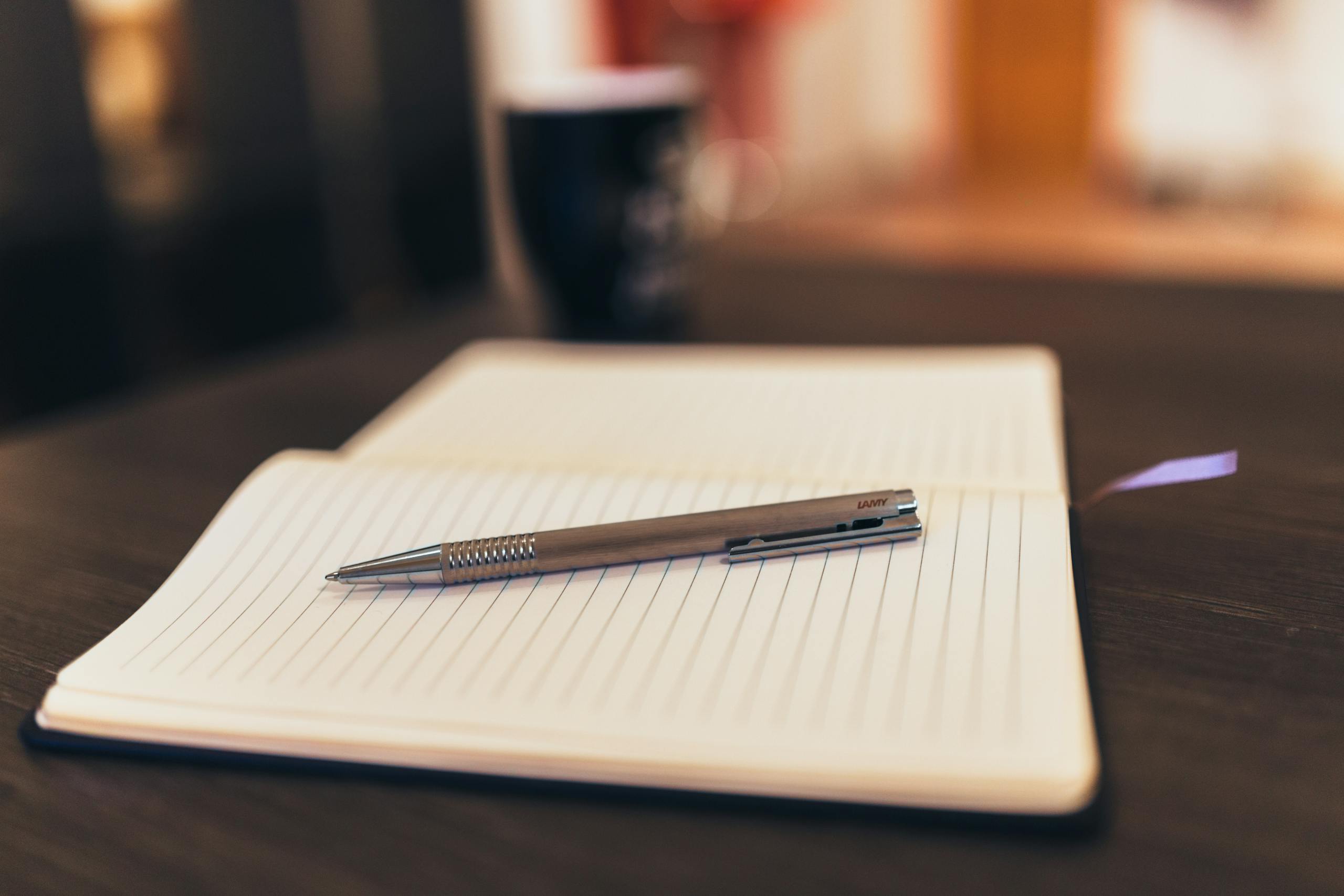 Open notebook with pen on a table, mug in background, cozy setting.